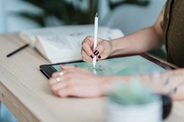 Designer holding a white stylus pen while sketching on a tablet displaying soft blue-green watercolor tones on a wooden work surface, with a notebook and blurred materials in the background, conveying a focused and creative design process